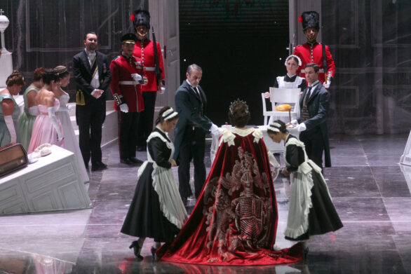 Degli attori sono in scena sul palco durante l'opera "Elisabetta Regina d'Inghilterra". La regina è girata di spalle e indossa un mantello rosso; ai suoi lati si trovano le due cameriere. Nello sfondo altri personaggi