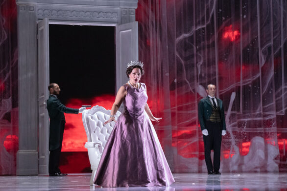 Degli attori sono in scena sul palco durante l'opera "Elisabetta Regina d'Inghilterra". La regina, in abito viola, sta cantando al centro del palco. Sullo sfondo le guardie