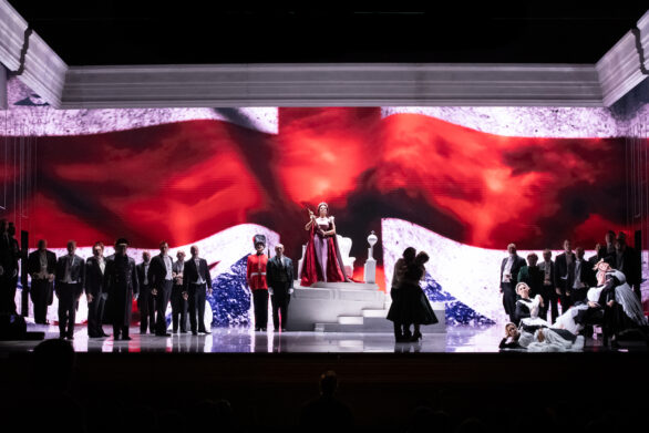 Degli attori sono in scena sul palco durante l'opera "Elisabetta Regina d'Inghilterra". La regina si trova su un palchetto bianco e indossa la corona e un mantello rosso. Ai suoi lati si trovano altri personaggi.